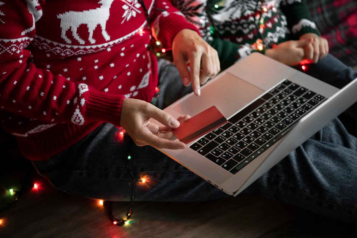 Homme avec pull de Noel et carte bleue dans la main et PC sur les genoux, ravi des baisses de prix des abonnements Internet, constatées par le baromètre des prix de décembre